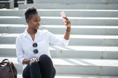 Beyaz merdivenlerde otururken akıllı telefon kullanan Afrikalı kadın..