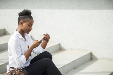 Beyaz merdivenlerde otururken akıllı telefon kullanan Afrikalı kadın..