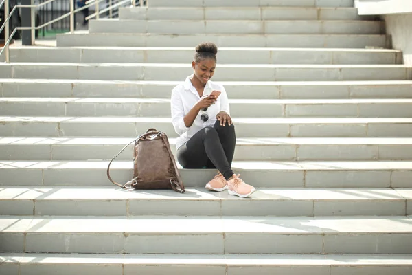 Africká žena pomocí chytrého telefonu, zatímco sedí na bílých schodech venku. — Stock fotografie