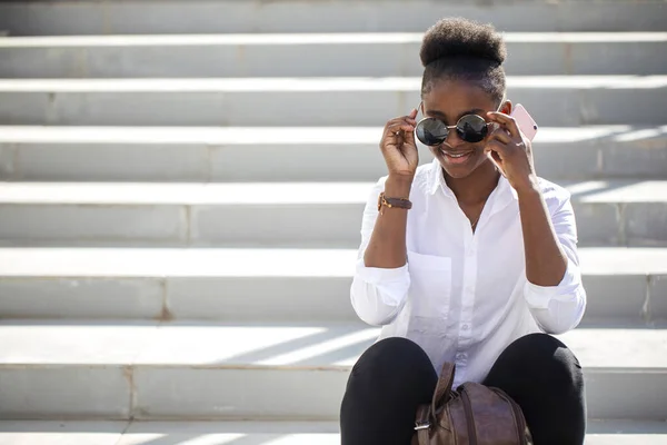 Afrikanerin benutzt Smartphone, während sie auf weißen Treppen im Freien sitzt. — Stockfoto