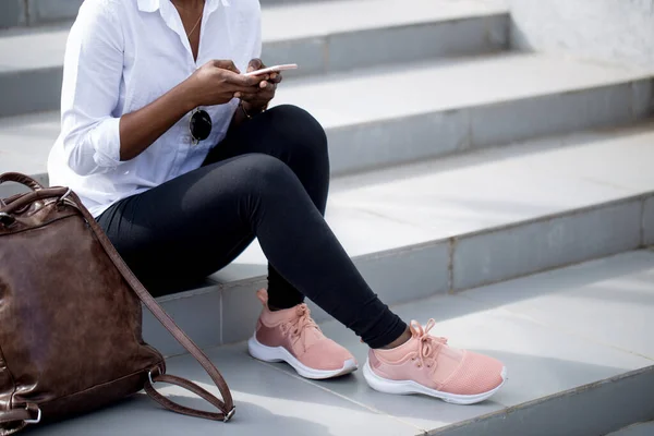 Afrikaanse vrouw met behulp van smart phone terwijl zitten op witte trappen buiten. — Stockfoto