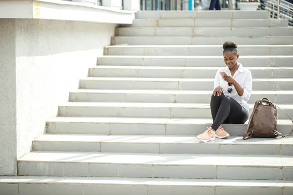 Donna africana utilizzando smart phone mentre seduto su scale bianche all'aperto. — Foto Stock