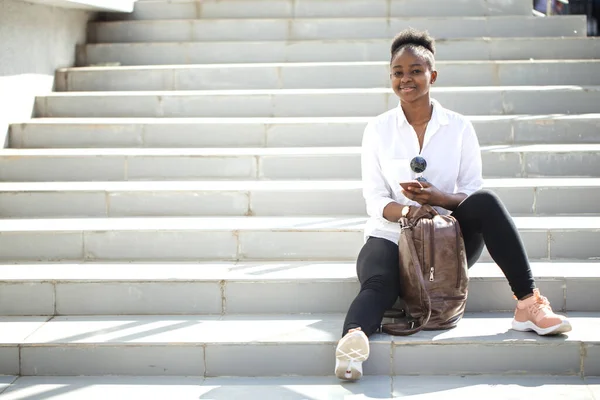 Mujer africana usando teléfono inteligente mientras está sentado en escaleras blancas al aire libre. —  Fotos de Stock