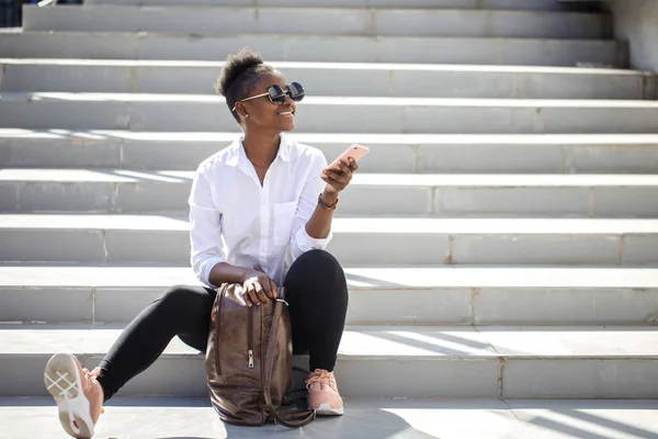 Mujer africana usando teléfono inteligente mientras está sentado en escaleras blancas al aire libre. —  Fotos de Stock