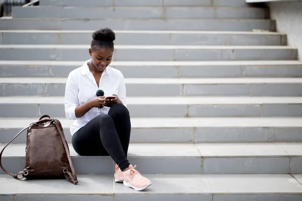 Donna africana utilizzando smart phone mentre seduto su scale bianche all'aperto. — Foto Stock