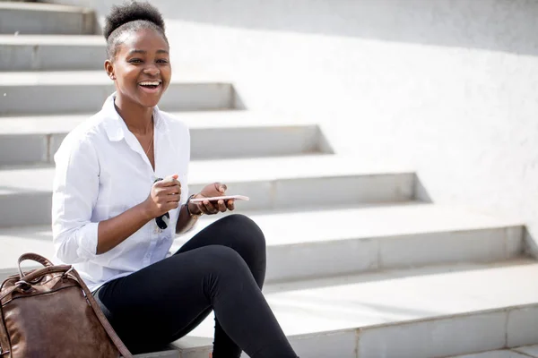 Afrikansk kvinna som använder smart telefon medan du sitter på vita trappor utomhus. — Stockfoto