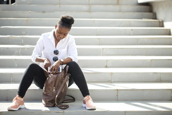 Donna africana utilizzando smart phone mentre seduto su scale bianche all'aperto. — Foto Stock