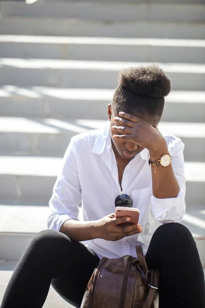 Afrikanerin benutzt Smartphone, während sie auf weißen Treppen im Freien sitzt. — Stockfoto
