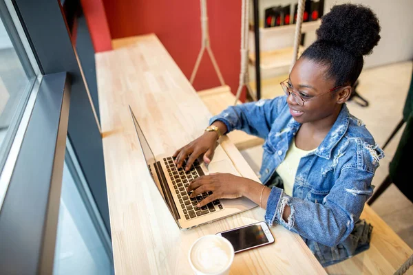 Mulher africana em jaqueta jeans sentado perto da janela e usando laptop no café — Fotografia de Stock