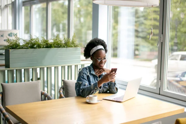 Young African woman with frizzy dark hair shopping online while sitting in cafe.