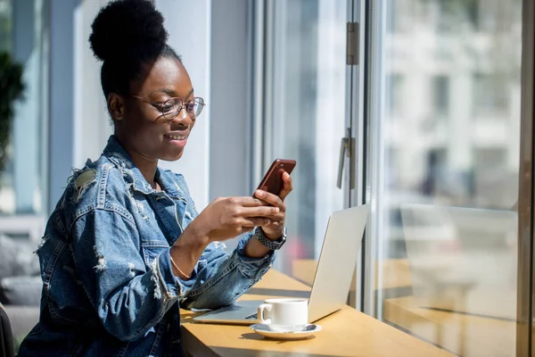 Hipster mujer de piel oscura con peinado afro hablando en el teléfono inteligente en la cafetería. — Foto de Stock