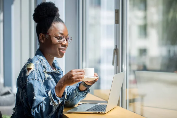 Afrikansk kvinna i denim jacka sitter nära fönstret och använder laptop i café — Stockfoto