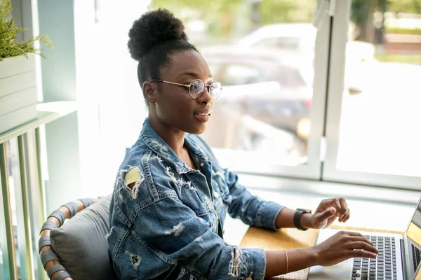 Afričanka v džínové bundě sedí u okna a pomocí notebooku v kavárně — Stock fotografie