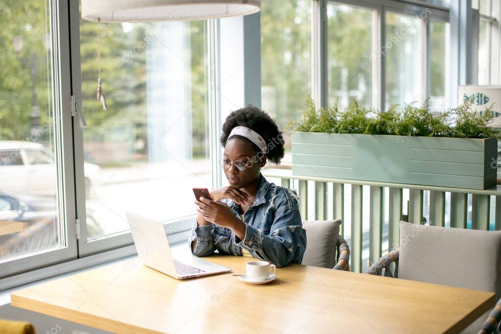 Young African woman with frizzy dark hair shopping online while sitting in cafe.