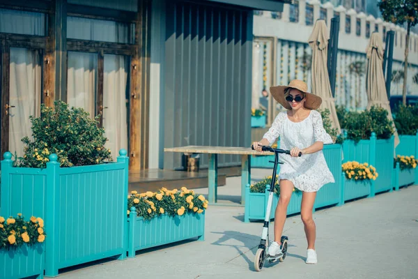 하얀 드레스를 입고 모자를 쓴 금발 여자가 파란 킥 스쿠터를 타고 도로를 달리고 있다 — 스톡 사진