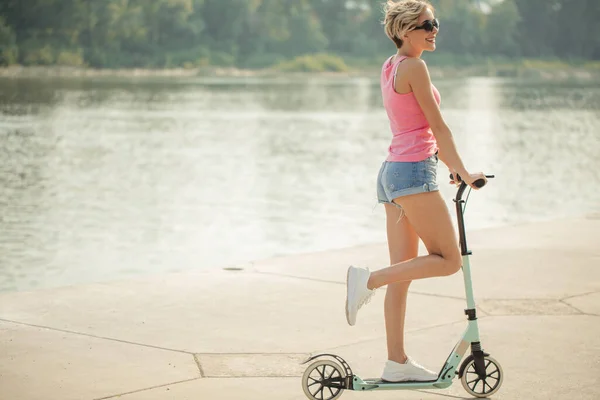 Bonito feminino piloto em sapatos brancos está montando no azul chute scooter na estrada no parque no dia ensolarado — Fotografia de Stock