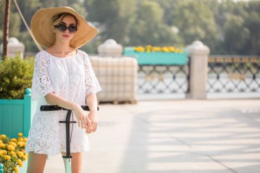 Modayı takip eden kadın portresi, beyaz elbiseli güzel sarışın kız, güneş gözlüğü ve scooter süren şapka.