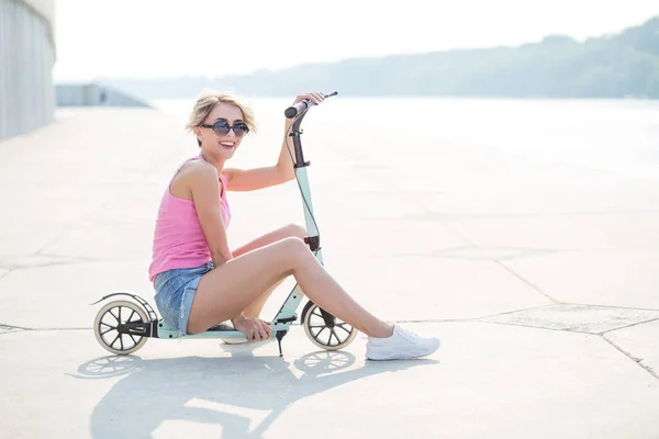 Femme blonde positive assise au scooter coup de pied bleu — Photo