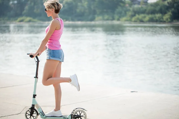 Schattig vrouwelijk rijder in witte schoenen is paardrijden op blauwe kick scooter op de weg in het park op zonnige dag — Stockfoto