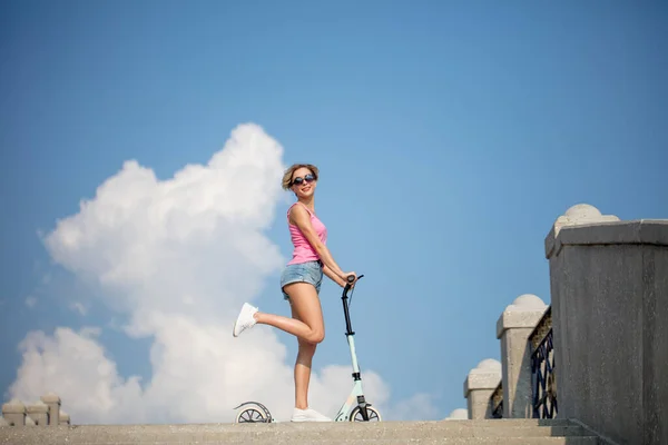 Blond kvinna poserar med blå sparkcykel på himlen bakgrund — Stockfoto