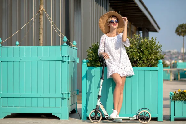 Vrouw in witte jurk en hoed poseren op blauwe kick scooter op de weg op zonnige dag — Stockfoto