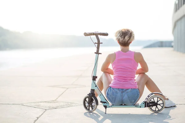 Mulher loira positiva sentada na scooter pontapé azul — Fotografia de Stock