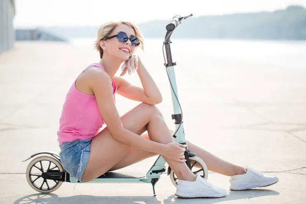 파란 킥 스쿠터에 앉아 있는 긍정적 인 금발 여성 — 스톡 사진