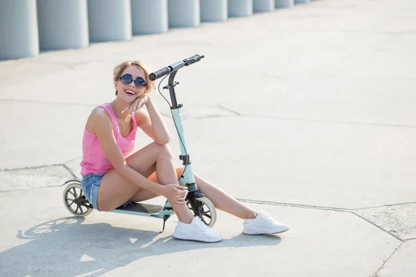 Femme blonde positive assise au scooter coup de pied bleu — Photo