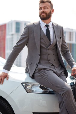 İyi görünümlü sakallı iş adamı temsili araba kaputuna oturur.