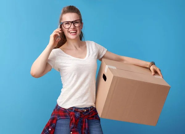 青い背景に携帯電話で話している段ボール箱と白いシャツで笑顔の若い女性 — ストック写真