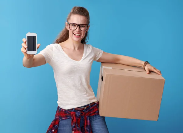 青の背景に携帯電話の画面を見せて 段ボール箱と白いシャツの若い女性を笑顔 — ストック写真