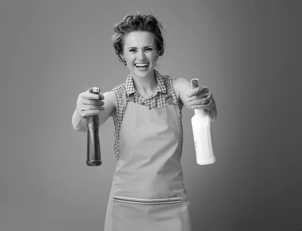 愉快的现代家庭主妇在围裙使用清洁洗涤剂的瓶作为枪 — 图库照片