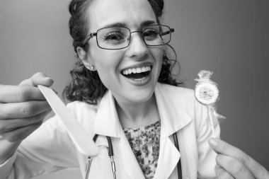 Boğaz arka plan üzerinde izole incelemek için bir spatula kullanarak lolipop ile beyaz tıbbi elbise içinde gülümseyen pediatrist doktor