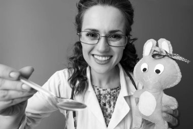 Çocuk doktoru arka plan üzerinde çocuk süspansiyon dikkat dağıtıcı çocuk oyuncağı ile süre veren beyaz tıbbi elbise içinde gülümseyen