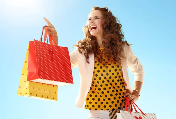 青空を指してショッピング バッグと薄手のジャケットでスタイリッシュな女性の幸せ — ストック写真
