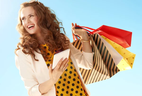 微笑时尚的女人穿着轻便夹克与平板电脑的购物袋 看着蓝天的一边 — 图库照片