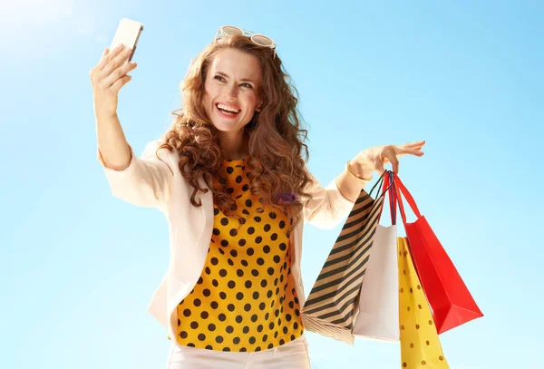 青空の携帯電話で Selfie を取って買い物袋と薄手のジャケットで幸せなトレンディな女 — ストック写真