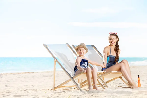 happy modern mother and daughter in swimsuit on seashore sitting on beach chairs