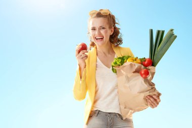 elma mavi gökyüzü karşı ısırma yiyecek ile kese kağıdı ile sarı ceketli mutlu modern kadın