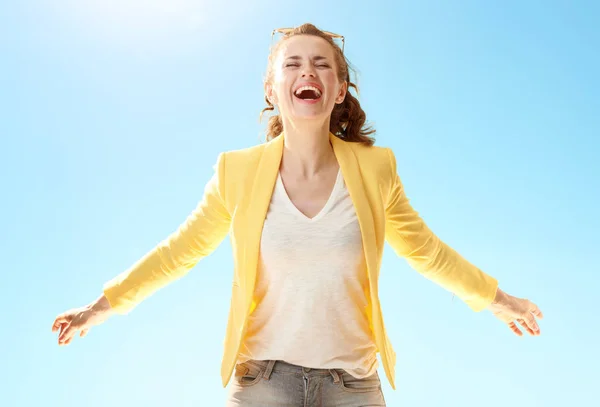 smiling modern woman in yellow jacket rejoicing against blue sky