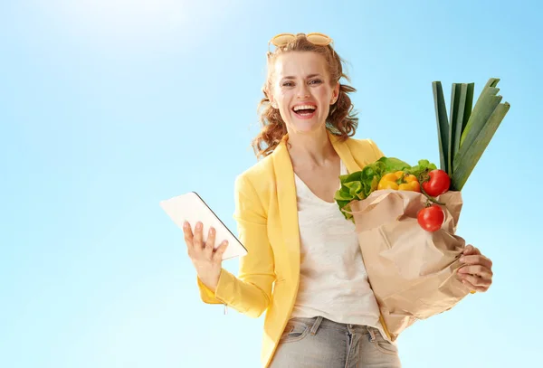 微笑时尚的女人穿着黄色夹克与纸袋与食品杂货与平板电脑对蓝天 — 图库照片