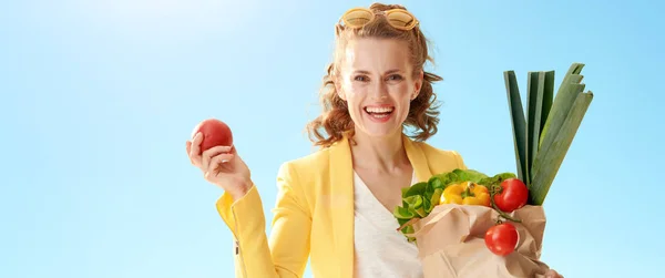 微笑的年轻妇女在黄色夹克用纸袋子与杂货拿着苹果对蓝天 — 图库照片