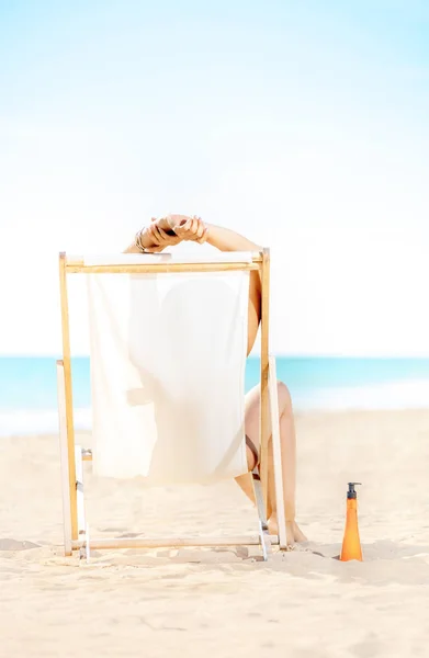 ビーチチェアに座っている日焼け止めローションを海岸で水着の現代の女性の後ろから見てください — ストック写真