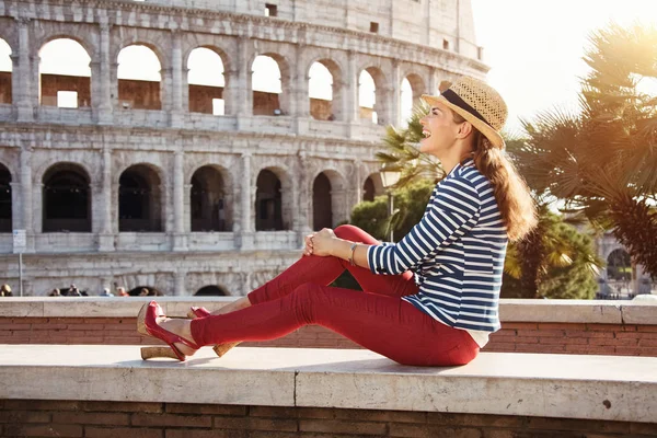 happy trendy woman in a striped jacket in the front of Colosseum looking into the distance while sitting