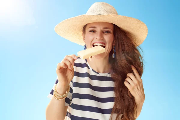 微笑的年轻妇女在草帽反对蓝色天空咬冰淇淋 — 图库照片