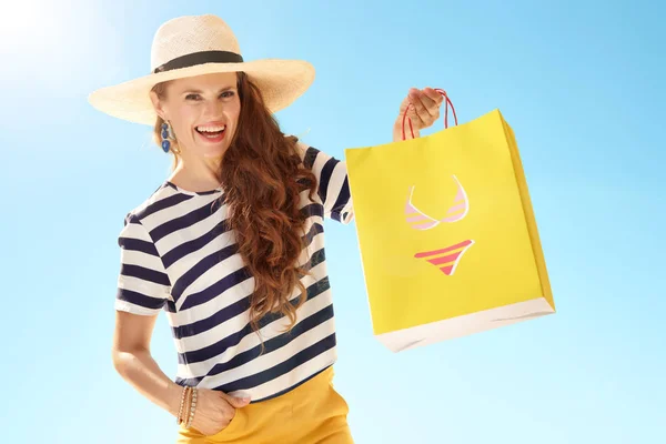 麦わら帽子ビキニで黄色の買い物袋を示す青い空を背景に現代女性の笑みを浮かべてください — ストック写真