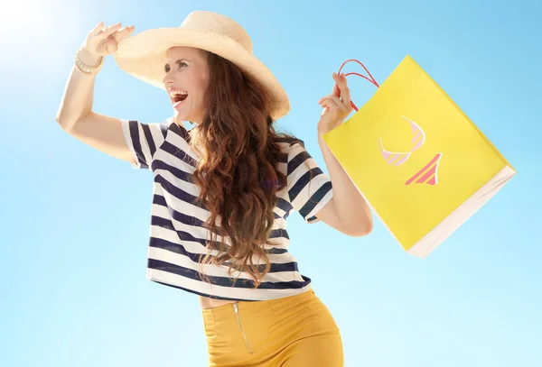 麦わら帽子の距離にビキニで黄色のショッピング バッグと青い空を背景に幸せなトレンディな女 — ストック写真