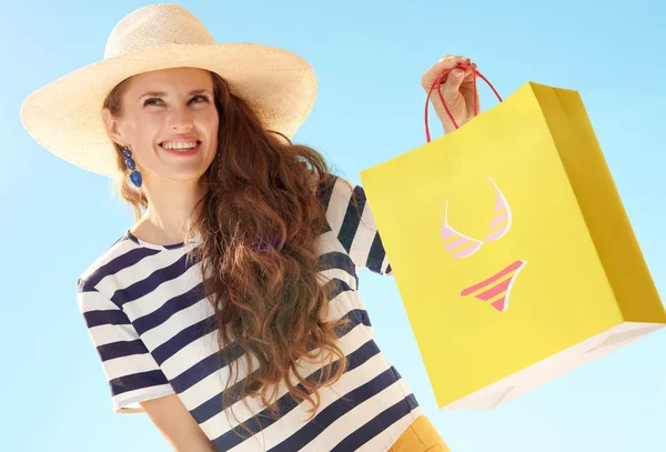 麦わら帽子ビキニで黄色の買い物袋と青い空を背景に物思いにふける現代女性 — ストック写真