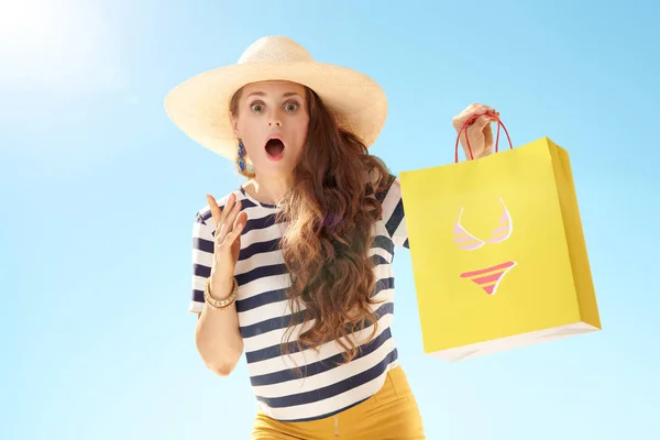 麦わら帽子ビキニで黄色の買い物袋と青い空を背景に驚くトレンディな女性 — ストック写真