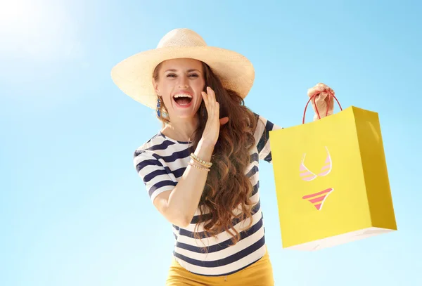 エキサイティングなニュースを伝えるビキニで黄色の買い物袋と青い空を背景に麦藁帽子で幸せなトレンディな女 — ストック写真